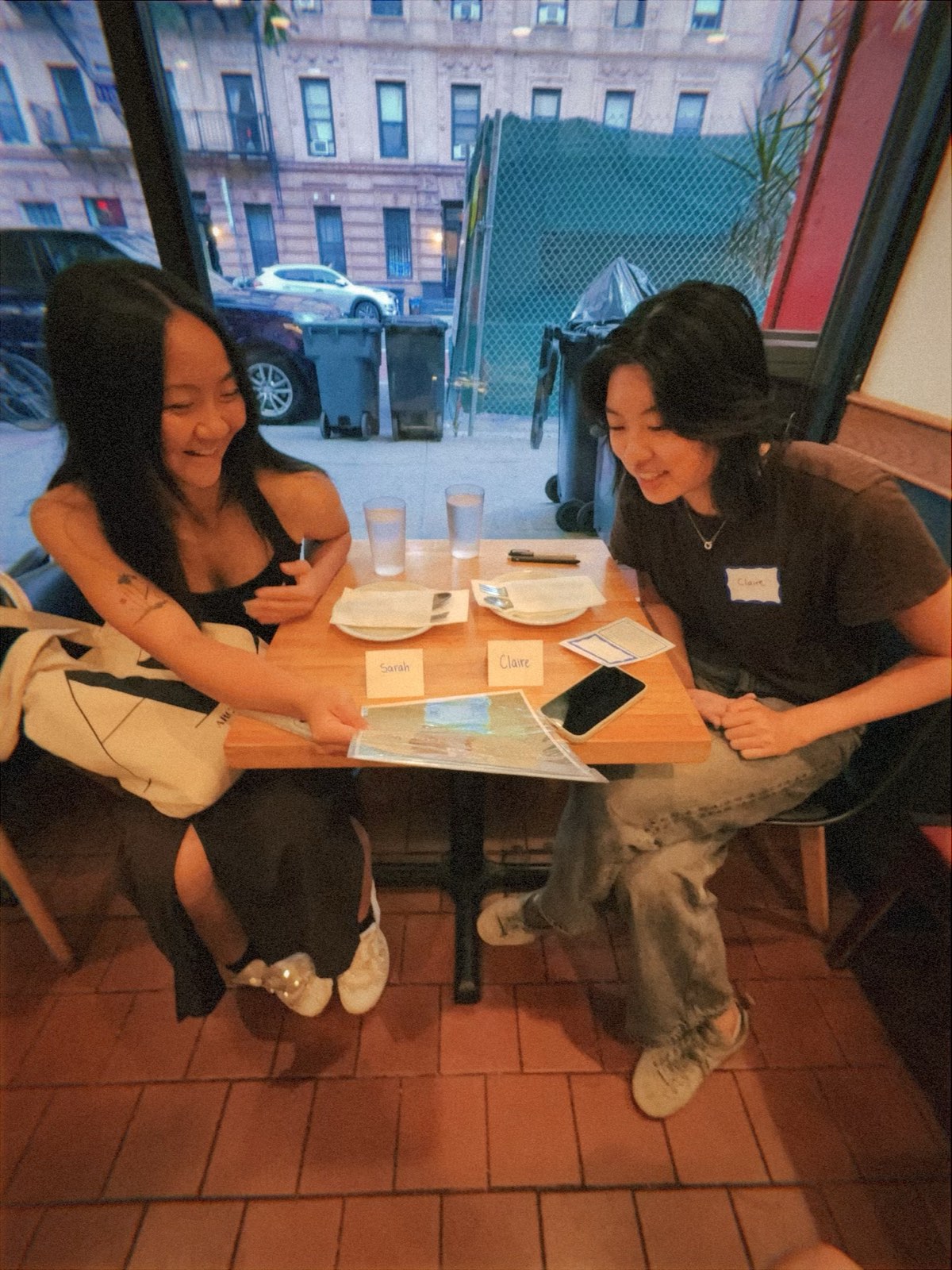 Two attendees sitting across a small table by the window, looking down at a menu together and laughing. Handwritten name cards sit on the table between them.