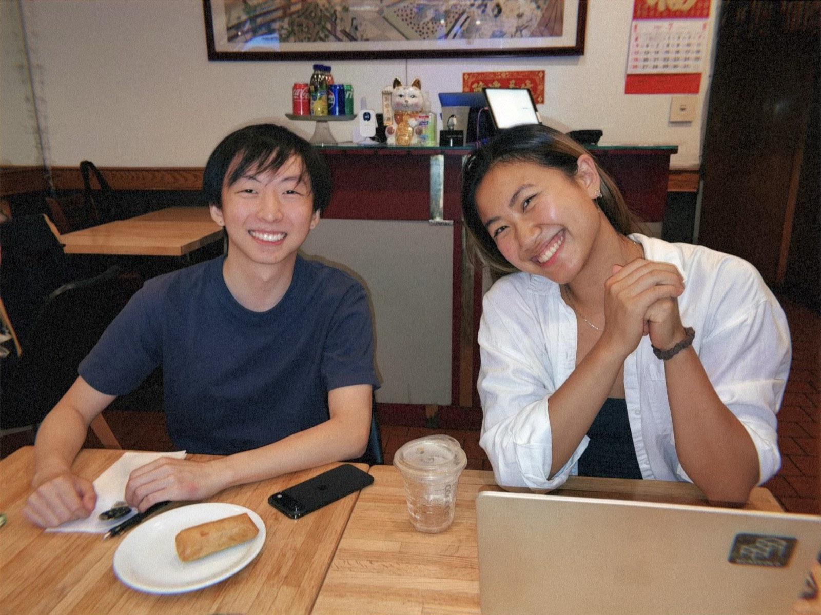 Two of the Scaffold team seated at a table, smiling at the camera, with a laptop, an iced drink and a plate holding a veggie roll in front of them.