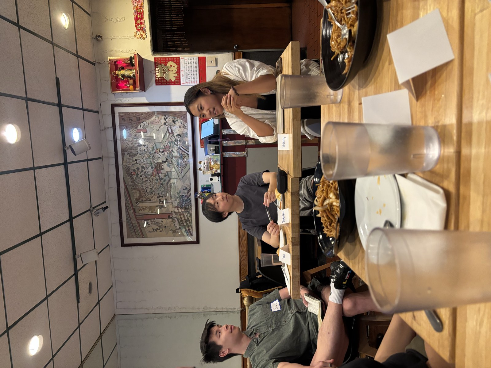 The intro panel underway — three people seated at the front of the restaurant in conversation, with name cards reading Sam, Brendan and Hillary on the table, and bowls of lo mein in the foreground.